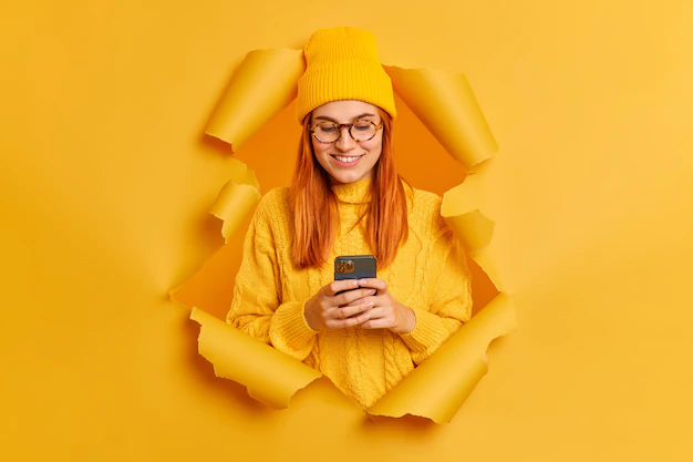beautiful-redhead-woman-uses-mobile-phone-surfs-social-networks-has-good-mood-dressed-yellow-hat-sweater-breaks-through-paper-hole_273609-46612
